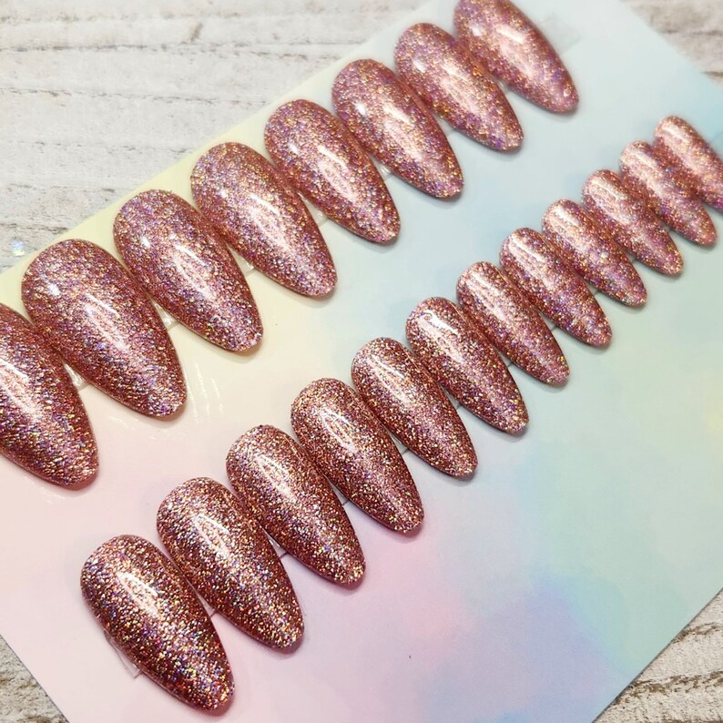 May include: Rows of rose gold glitter press-on nails in various sizes and shapes. The nails have a glossy finish and are arranged on a light-colored surface. The glitter reflects light, creating a sparkling effect. The nails are designed for cosmetic use.