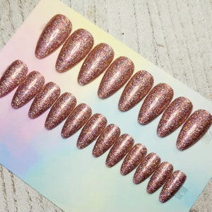 May include: Rows of rose gold glitter press-on nails in various sizes and shapes. The nails are oval-shaped and covered in sparkling glitter. The nails are arranged on a light-colored surface, ready for application.