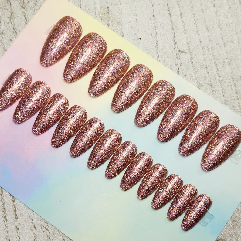 May include: Rows of rose gold glitter press-on nails in various sizes and shapes. The nails are oval-shaped and covered in sparkling glitter. The nails are arranged on a light-colored surface, ready for application.