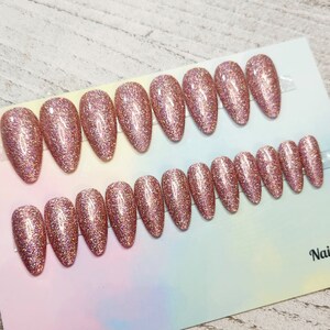 May include: A set of rose gold glitter press-on nails in various sizes. The nails are oval-shaped and arranged on a white and pastel-colored backing. The glitter effect gives a sparkling appearance, suitable for a glamorous look.