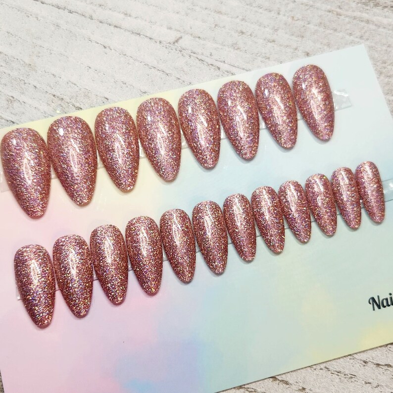 May include: A set of rose gold glitter press-on nails in various sizes. The nails are oval-shaped and arranged on a white and pastel-colored backing. The glitter effect gives a sparkling appearance, suitable for a glamorous look.