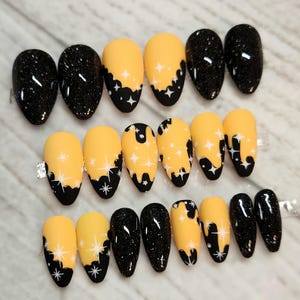 May include: A set of press-on nails with a variety of designs. Some nails are solid black with glitter, while others are yellow with black dripping designs and white star accents. The nails are oval-shaped.