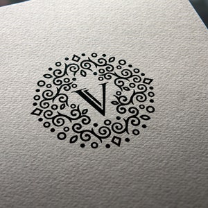 May include: A black and white monogram design featuring the letter "V" surrounded by a decorative circular frame of swirls and dots.