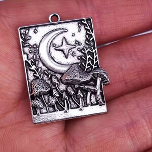 May include: A silver-toned rectangular pendant featuring a crescent moon, a star, and several mushrooms. The pendant has a bail at the top for attaching to a chain. The design includes detailed foliage and a textured background.