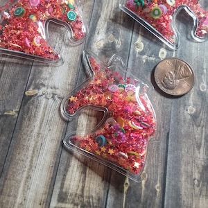 May include: Clear plastic unicorn head-shaped shaker filled with red star-shaped glitter and fruit-shaped confetti. A penny is next to the shakers. The shakers are on a wooden surface.