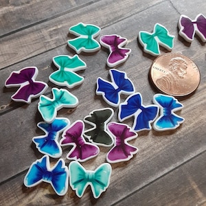 May include: A collection of colorful bow-shaped embellishments in shades of teal, purple, and blue, with white outlines. A penny is included for size reference. The bows are scattered on a wooden surface.