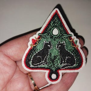 May include: Embroidered patch featuring two black cats facing each other within a green and white design. The patch has a red border and a white backing. The design is in the shape of a stylized tree.