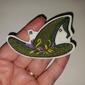 May include: Embroidered witch hat patch with a green and gold design. The hat features a pointed crown and a wide brim, with purple and yellow floral accents. The patch has a white border.