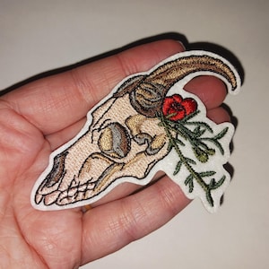 May include: Embroidered patch depicting a goat skull with brown horns and a beige skull. A red flower and green leaves are on the right. The patch has a white border, suitable for clothing or accessories.
