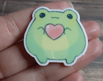 May include: A green frog holding a pink heart in its hands. The frog is cartoon-style and has a happy expression.