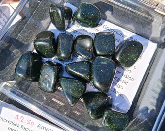 Puede incluir: Una colección de piedras preciosas de goldstone negro pulido con inclusiones verdes brillantes. Las piedras tienen varias formas y tamaños, exhibidas en un recipiente de plástico transparente. Una etiqueta blanca con texto es parcialmente visible.