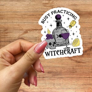 May include: A die-cut sticker featuring the text BUSY PRACTICING WITCHCRAFT, a skull with a purple candle, a glass bottle, a wine glass filled with purple liquid, a yellow shaker, and a lemon slice, all accented with small black star symbols.