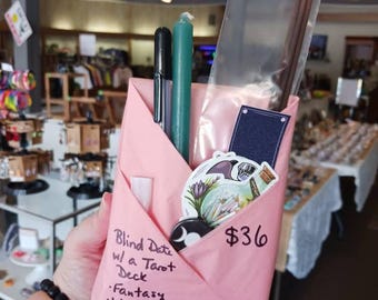 May include: A pink paper envelope containing a black pen, a teal candle, a clear plastic bag with brown incense sticks, a dark blue rectangular card, a sticker of a potion bottle with flowers, and a small black button with a white moon symbol.