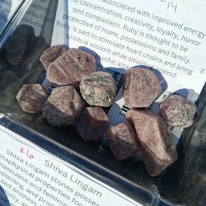 May include: A collection of rough, reddish-brown Shiva Lingam stones. The stones are displayed in a clear container with a price tag and a description of their metaphysical properties. The text on the tag mentions awakening and creativity.