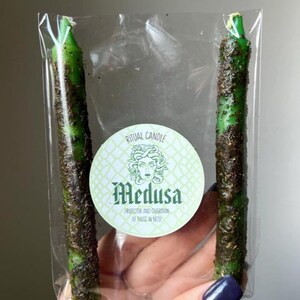 May include: Two green taper candles coated in dark herbs, packaged in a clear plastic sleeve. A circular label in the centre features a green Medusa illustration, the text RITUAL CANDLE, Medusa, and PROTECTOR AND GUARDIAN OF THOSE IN NEED.
