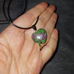 Puede incluir: Collar con colgante en forma de corazón con una piedra verde y morada. El colgante cuelga de un cordón negro. La piedra tiene una superficie pulida y está engastada en un metal plateado. El collar se muestra en una mano.