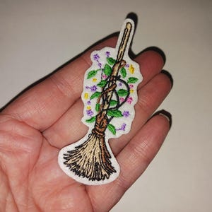 May include: Embroidered patch featuring a broom with a wooden handle and straw bristles. The broom is adorned with green leaves, purple and yellow flowers, and a black pentacle symbol. The patch is on a white background.
