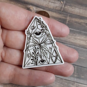 Puede incluir: Una pegatina blanca con una ilustración en blanco y negro de una pirámide geométrica con dos ojos y diseños florales.
