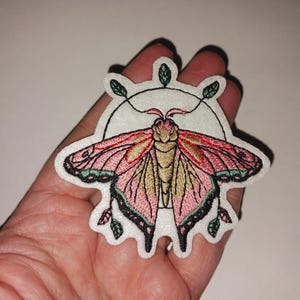 May include: Embroidered moth patch with pink, gold, and black wings, surrounded by a white border. The moth design includes a circular element with leaves. The patch is held in a hand.