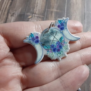 Puede incluir: Un colgante con diseño de media luna, un círculo central y una estrella de cinco puntas. Decorado con detalles florales azules y morados. Un anillo plateado está adjunto para colgarlo. Ideal como joya.