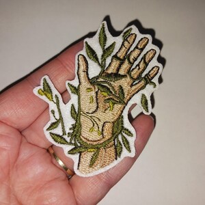 May include: Embroidered patch featuring a hand intertwined with green vines and leaves. The hand is tan with detailed stitching, and the vines have a natural green hue. The patch has a white border, suitable for application on clothing or accessories.