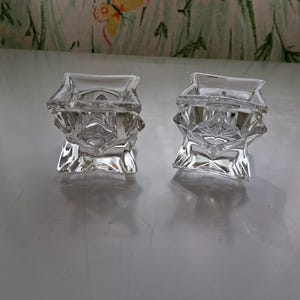 May include: Two clear glass votive candle holders featuring a geometric, faceted design with a square top opening and a sturdy base. The glass is transparent and reflects light, showcasing sharp angular edges and a multi-sided, crystalline structure.