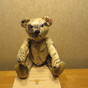 May include: A beige teddy bear with a newspaper print design. The bear has black eyes and a black nose. The bear is sitting on a white surface with a tag that says "Steiff Club".