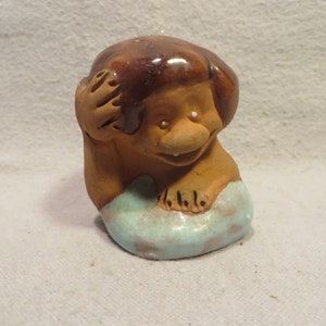 May include: A small ceramic figurine of a brown-haired person with a toothy grin, sitting on a blue and brown base.