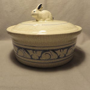 May include: A round ceramic lidded casserole dish with a cream-colored crackle glaze finish. The lid features a sculpted rabbit figurine as a handle. The base of the dish is decorated with a dark blue band containing a repeating leaf pattern.