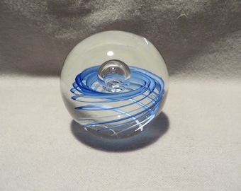 May include: A clear glass paperweight with a blue and white swirl design inside. The swirl is centered around a smaller clear glass sphere.