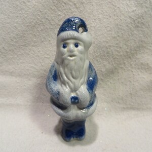 May include: Blue and white ceramic Santa Claus ornament. The Santa is wearing a blue hat and coat with white trim. He has a white beard and a white face with blue eyes.