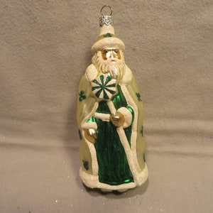 May include: A glass Christmas ornament depicting a Santa Claus figure wearing a white fur-trimmed robe with green shamrock accents. He holds a candy cane with a shamrock design.