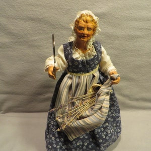 May include: A vintage doll wearing a blue and white dress with a floral pattern. The doll is holding a bundle of lavender and a small metal tool.