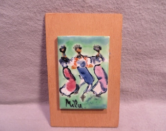 May include: A framed ceramic tile with a green background and three figures in pink, blue, and white dresses dancing. The tile is signed "Mila" in the bottom left corner.
