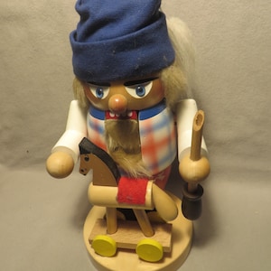 May include: A wooden nutcracker figurine with a blue knit cap, white hair, and a plaid vest. The nutcracker holds a wooden pipe and a toy horse with a red saddle. The base is round and made of light-colored wood.