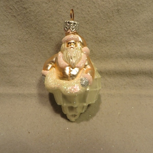 May include: A glass Christmas ornament featuring a Santa Claus figure in a pink and gold suit, sitting in a white, glittery sleigh.