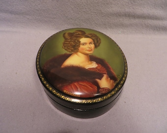 May include: A round black lacquered wooden box with a painted portrait of a woman with dark hair and a fur collar. The box is decorated with gold trim and the word "FEANKINO" is visible on the bottom.