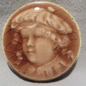 May include: A round ceramic tile with a raised image of a woman's face. The tile is brown with a crackled glaze and a white rim.