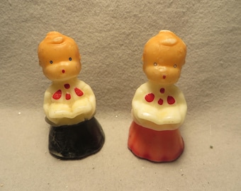 May include: Two molded wax figurines depicting children with peach-colored skin, light hair, and painted facial features. Each figure wears a white top with red accents and holds an open book. One figure has a black base, while the other has a red base.