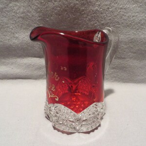 May include: A red glass pitcher with a clear glass handle. The pitcher has a clear glass base with a decorative pattern. The pitcher has gold lettering that says "New 1922  P.S."