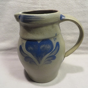 May include: A gray stoneware pitcher with a blue heart and floral design. The pitcher has a wide mouth and a handle.