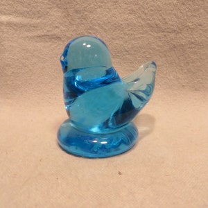 May include: A small, blue glass bird figurine. The bird is sitting on a base and has a rounded body and a small tail.