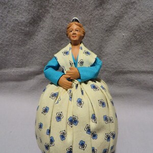 May include: A ceramic doll wearing a blue and white dress with a floral pattern. The doll has a white cap and brown hair.