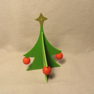 May include: A green, three-dimensional wooden Christmas tree ornament featuring a gold-colored star at the top and three small, round, red wooden balls attached to the branches as decorative elements. The structure is composed of intersecting flat panels.