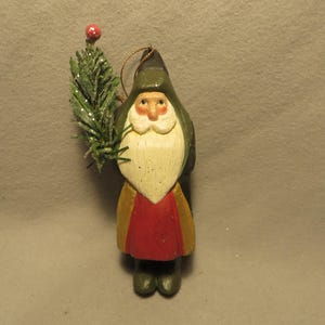 May include: A hand-painted wooden Santa Claus ornament wearing a dark green hooded cloak, a red tunic, and dark green boots. The figure has a long white beard and holds a small, artificial green pine branch topped with a single red berry.