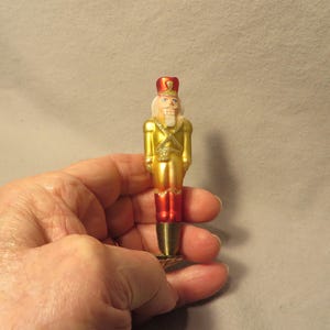 May include: A glass nutcracker ornament featuring a shiny gold uniform with red accents on the hat and legs. The figure has a painted face with a white beard and a metallic, ribbed conical base at the bottom, designed for display or attachment.