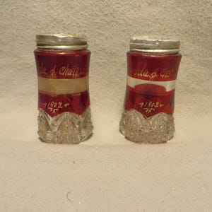 May include: Two vintage glass salt shakers with red glass and silver tops. The shakers have a decorative pattern and are inscribed with the text "Ida J. Chatten" and "1/1903" and "Ida J. Chatten" and "1/1903".