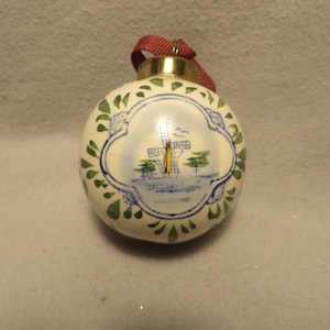 May include: A round, cream-colored ceramic ornament featuring a hand-painted blue and green windmill scene within a central cartouche. The ornament has a gold-toned metal cap at the top and a red fabric ribbon loop for hanging purposes.