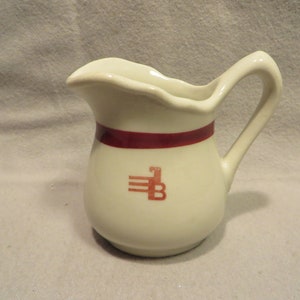May include: White ceramic creamer with a maroon band and a red logo of a stylized "B" with three lines above it.