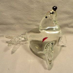 May include: A clear glass sculpture of a seal with a black glass ball on top of its head. The seal is lying on its side with its flippers outstretched. There is a small red glass piece inside the sculpture.